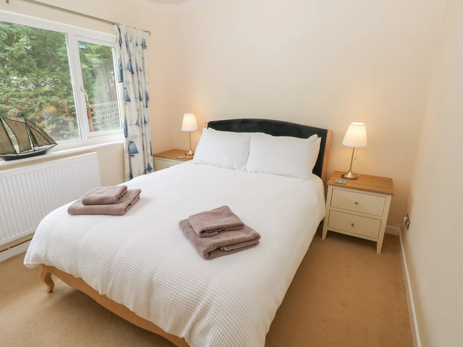 A downstairs bedroom with a king-size bed and bedsize drawers on each side, both with a lamp on top. There is also a large window and radiator.