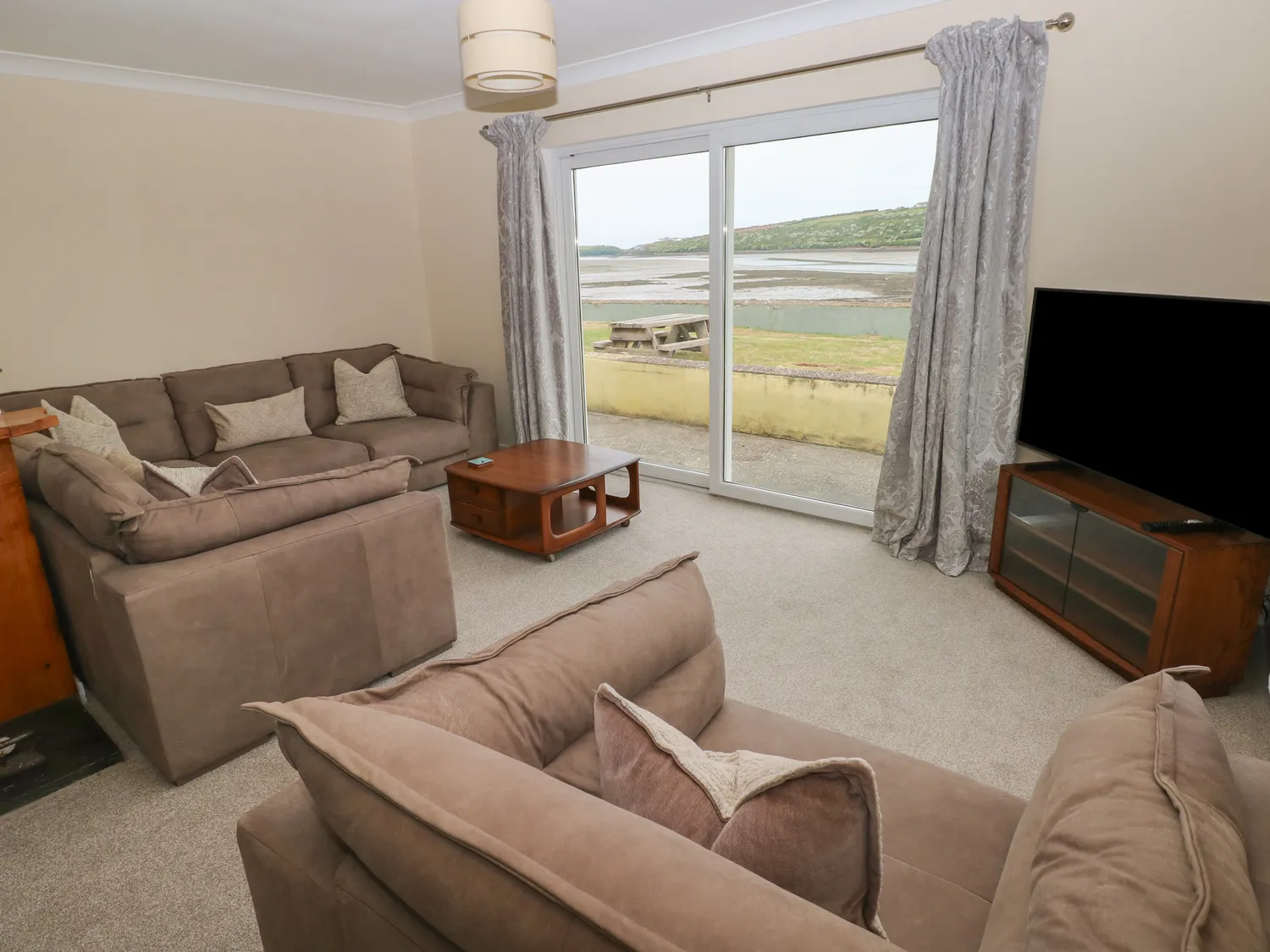Two large sofas and a large chair around a coffee table. They face large bay windows which give a view of the garden and river. There is also a large flatscreen tv on top of a cabinet in the corner.