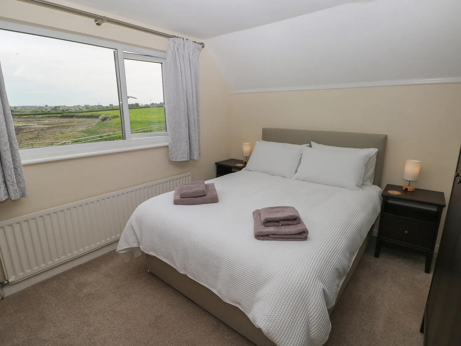 An upstairs bedroom with a king-size bed and bedsize drawers on each side, both with a lamp on top. There is also a large window and radiator.